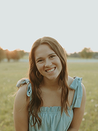 A woman in a light blue blouse poses in front of a green field with trees in the distance.