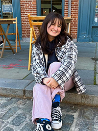 A young woman smiles while sitting casually on the sidewalk, dressed in a checked jacket and pink pants, with chairs and a café entrance in the background.