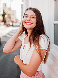 Smiling young woman wearing a white short-sleeve top, standing outdoors with soft sunlight illuminating her face.