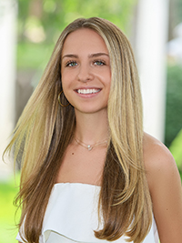 A young woman with long, straight blonde hair smiles warmly while wearing a white top, set against a blurred green background.