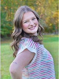 Smiling young woman with wavy hair wearing a colorful knit top, posing outdoors in a grassy setting.