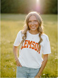 A young woman stands in a sunlit field wearing a white Clemson University t-shirt, smiling warmly at the camera.
