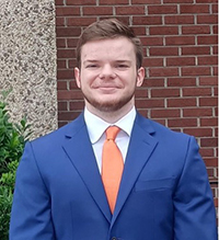 A young man in a blue suit with an orange tie smiling in front of a brick wall.