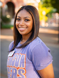 A smiling student wearing a purple Clemson University t-shirt stands outdoors in a sunny setting.
