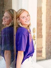 A smiling young woman in a purple t-shirt leans against a door with a reflective surface.