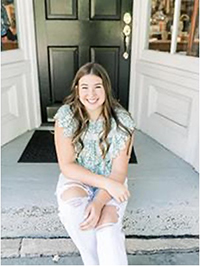 A young woman smiling while sitting on steps in front of a black door, wearing a stylish top and ripped jeans.