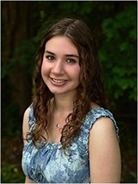 A young woman with curly hair smiles while wearing a blue floral dress, set against a green natural background.