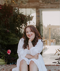 A woman sitting gracefully among vibrant greenery in a sunlit space, smiling and resting her chin on her hand.