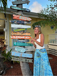 A young woman stands next to a colorful directional signpost with various locations marked, wearing a white top and a blue skirt.