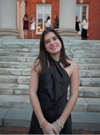 A young woman poses in a black dress on a set of stone steps. In the background, a group of people is gathered near a building.