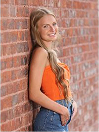 A young woman in an orange top leans against a brick wall, smiling confidently.