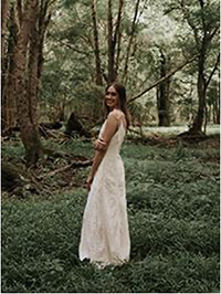 A woman in a white wedding dress stands gracefully in a forest surrounded by lush greenery.