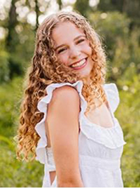 A young woman with curly hair smiles brightly while wearing a white dress with ruffled sleeves, set against a lush green background.