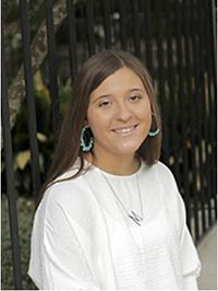 A smiling young woman with long brown hair wearing a white sweater and turquoise earrings, posing in front of a black railing.