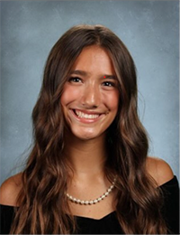 A portrait of a smiling young woman with long, wavy hair wearing an off-the-shoulder top and a pearl necklace.