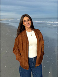 A young woman smiling on the beach, wearing a brown jacket and standing on wet sand with gentle waves in the background.