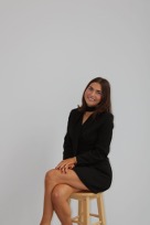 A smiling woman in a black blazer seated on a stool against a plain background.