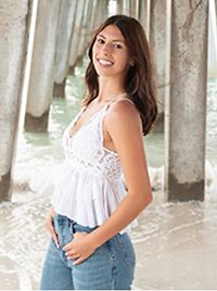 A young woman smiling while standing on the beach under a pier, wearing a white top and blue jeans.