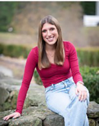 A smiling young woman in a red long-sleeve top and blue jeans sitting on a stone wall outdoors.