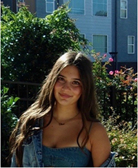 A young woman with long brown hair smiles while standing outdoors surrounded by greenery and flowers.