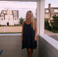 A woman in a dark dress stands on a porch, smiling with a seaside home visible in the background.
