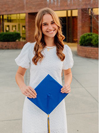 A smiling graduate in a white dress holds a blue graduation cap outside a school building.
