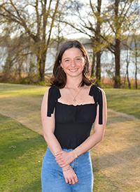 A young woman in a black top and jeans stands outdoors near a lake, smiling brightly.