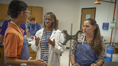 Students and a teacher engage in a science lab discussion while wearing safety goggles and lab coats.