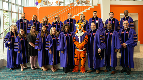 A group of Ph.D. graduates in dark purple robes stand in two rows with an orange Tiger mastcot in the middle front row.