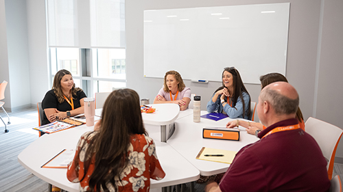 A group of five adults with orange lanyards sit around a white table in a bright and naturally lit classroom.