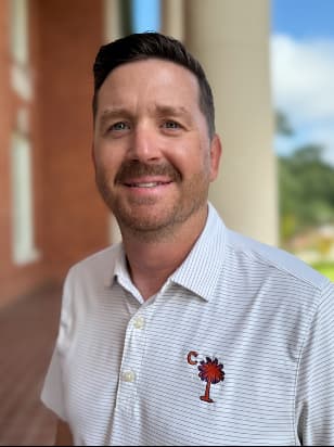 Smiling man in a Clemson University orange polo shirt sitting outdoors with greenery in the background.