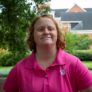Smiling woman with curly, reddish hair wearing a pink polo shirt stands outdoors, surrounded by greenery.