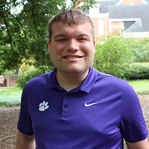A young man smiling outdoors in a purple polo shirt featuring a paw print logo.