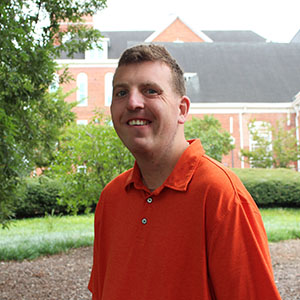 A smiling man in an orange shirt stands outdoors, surrounded by greenery and a brick building in the background.