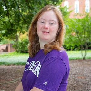 A young woman smiles while wearing a purple T-shirt in a lush outdoor setting.