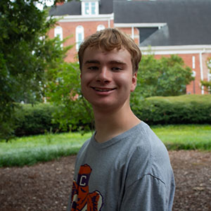 A young man smiles while sitting outdoors in a garden setting.