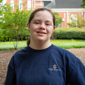 A smiling young woman wearing a navy blue t-shirt stands outdoors surrounded by greenery.