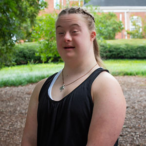 A young woman smiling outdoors, wearing a black tank top and a necklace, with greenery in the background.