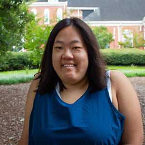 A young woman with dark hair smiles while wearing a blue tank top, set against a green garden background.