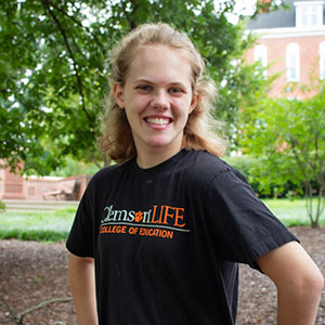 Smiling student wearing a black Clemson LIFE t-shirt stands outdoors with greenery in the background.