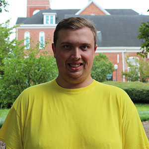 A young man smiles while sitting outdoors in a garden setting.