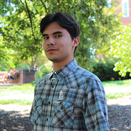 Young man in a plaid button-up shirt standing outdoors in a sunlit park.