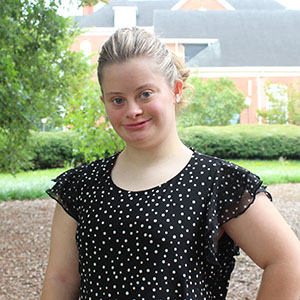 A young woman smiles confidently while wearing a black polka dot blouse in a green outdoor setting.