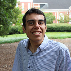 A smiling young man in a collared shirt stands outdoors with greenery in the background.