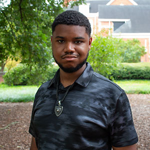 A young man with short hair and a beard wearing a dark polo shirt stands confidently outdoors.