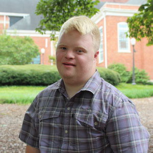 A young man smiles while sitting outdoors in a garden setting.
