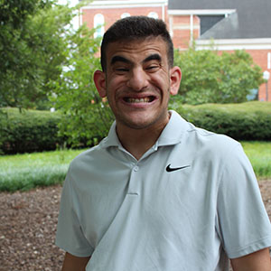 A young man smiles while sitting outdoors in a garden setting.