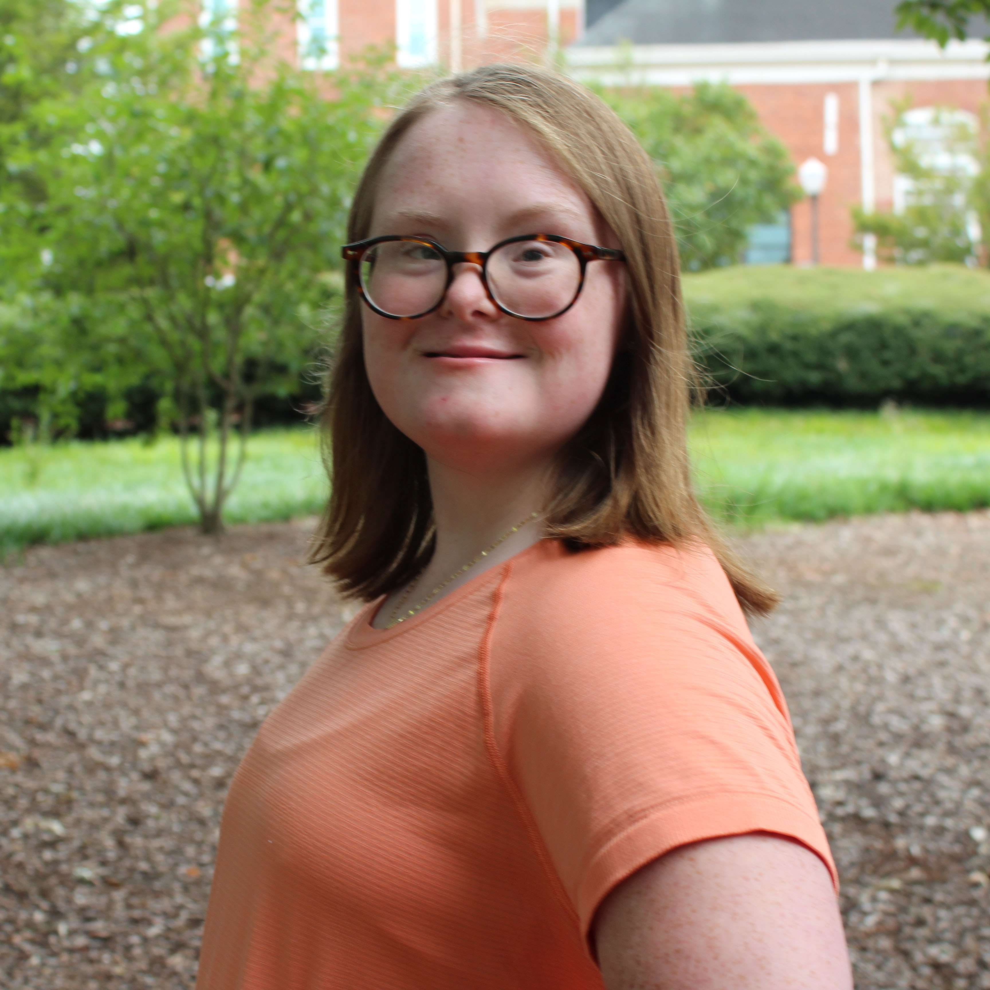 A smiling young woman with glasses and brown hair posing confidently outdoors in a park setting.