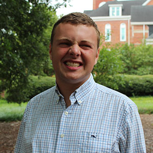 A smiling young man in a checkered shirt stands outdoors, surrounded by greenery and buildings in the background.