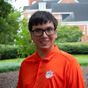 A young man wearing an orange polo shirt with a tiger paw logo, smiling against a background of greenery and a building.